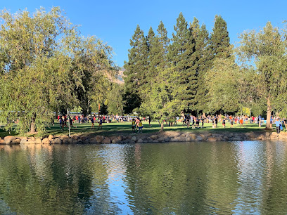 Park scene with river, green trees, and people gathering along the water on a sunny day