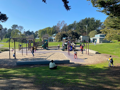 Community park with playground equipment, children playing, green lawn, and trees under blue sky